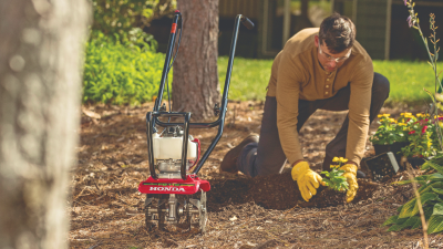 A gardener, on their knees, plants flowers behind a Honda tiller. 