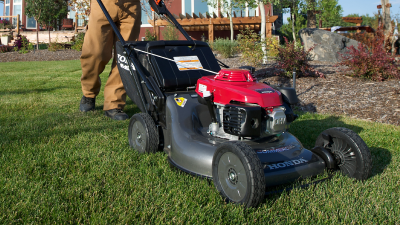 Un employé en entretien paysager pousse une tondeuse Honda près d’un buisson sur un terrain de golf. Un autre employé et un golfeur se trouvent en arrière-plan.