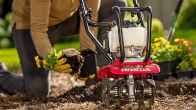 Gros plan d’un motoculteur dans un parterre de fleurs. En arrière-plan, un homme transplante des fleurs d’un pot au parterre.