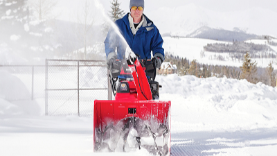 Image de face d'un homme en veste bleue poussant une souffleuse à neige Honda rouge. Paysage de neige en arrière-plan. Arbres et montagnes au loin.