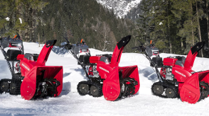 Wide view of three snowblowers parked side by side on a bed of snow with a coniferous forest in the background.  